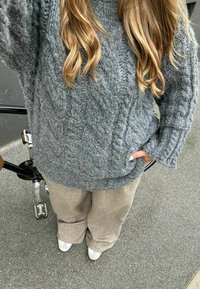 Maglione grigio lavorato a maglia con motivo a treccia, colletto alto e vestibilità morbida; abbinato a pantaloni beige a gamba larga e sneakers grigie su un terreno texturizzato.