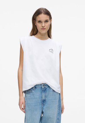 Ung kvinde iført en hvid ærmeløs top med polstrede skuldre og lyseblå løstsiddende jeans, stående mod en hvid baggrund.