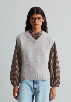 Junge Frau mit langen dunklen Haaren, die große braune Brille trägt, eine graue Strickweste über einer braun karierten Bluse und hellblaue Jeans.