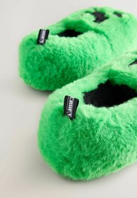 Chaussons en velours verts avec une texture pelucheuse, une forme arrondie et des accents noirs. Présente une étiquette portant la mention "CRAFT". Matériau doux et épais.