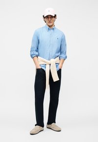 Άνδρας φορώντας γαλάζιο πουκάμισο με ρίγες και κουμπιά, μπεζ πουλόβερ δεμένο στη μέση, μαύρο παντελόνι, μπεζ μοκασίνια και ροζ καπέλο μπέιζμπολ.