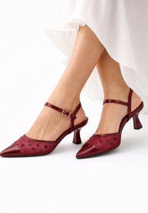 Frau trägt burgunderrote Slingback-Pumps mit spitz zulaufender Spitze, Knöchelriemen und gemusterten Mesh-Einsätzen, kombiniert mit einem weißen, fließenden Kleid.
