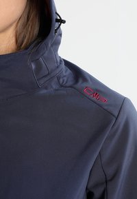 Woman wearing navy blue hooded jacket with red CMP logo embroidered on the shoulder, visible hood adjustment cord.