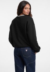 Donna che indossa una felpa nera con colletto a righe e jeans blu scuro con un logo triangolare sulla tasca posteriore, mentre guarda oltre la spalla.