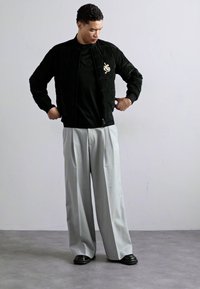 Sort quiltet bomberjakke med frontlynlås, båret over en sort skjorte. Grå brede bukser og sorte slip-on sko fuldender looket.