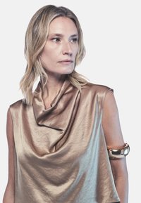 Top drappeggiato in bronzo con morbido lucido, caratterizzato da un collo a cowl e design senza maniche. Bracciale metallico che accentua lo stile.