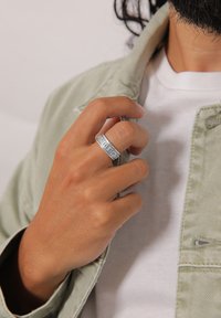 Silberner Ring mit erhabenen Buchstaben, an einer Hand getragen, im Hintergrund ist eine hellgrüne Jacke und ein weißes Hemd zu sehen. Glatte Oberflächenstruktur.