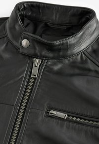 Jachetă din piele neagră, cu guler înalt, accesorii din metal argintiu, fermoar frontal și un buzunar cu fermoar pe piept, având un finisaj texturat.