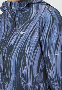 Gros plan d'une personne portant une veste Nike à fermeture éclair, avec un motif marbré bleu et noir, une capuche visible et le logo swoosh blanc sur la poitrine.