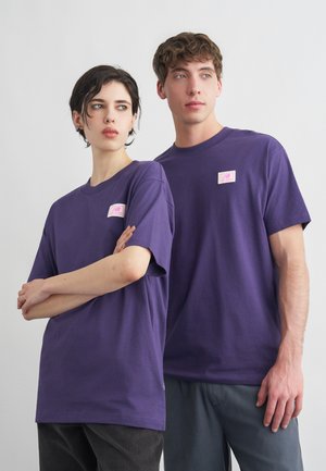 Δύο νέοι ενήλικες φοράνε όμοια μωβ μπλουζάκια με μικρά ροζ λογότυπα, στέκονται δίπλα δίπλα μπροστά σε ένα απλό φόντο.