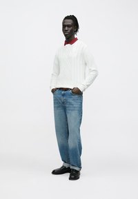 Biały sweter w splot warkoczowy założony na czerwoną koszulę z kołnierzykiem, do tego jasnoniebieskie jeansy o prostych nogawkach i czarne buty. Prosty, codzienny strój.