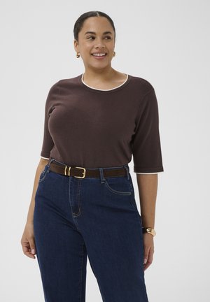 Femme souriante avec les cheveux attachés, portant des boucles d'oreilles créoles dorées, un haut marron à manches courtes, un jean bleu foncé avec une ceinture marron et un bracelet en or.