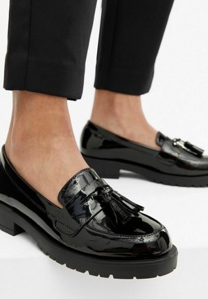Sorte laklæder loafers med kvaster, båret med sorte korte bukser på bare fødder mod hvid baggrund.