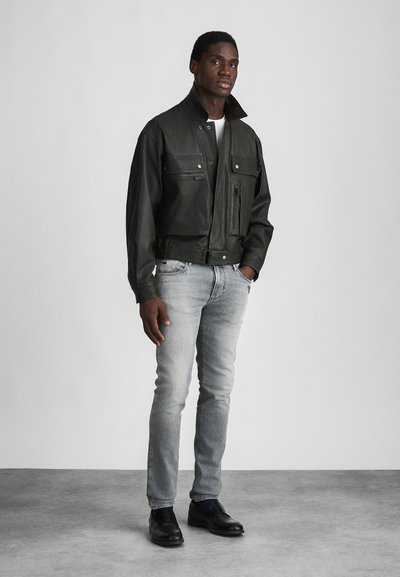 Πράσινο δερμάτινο bomber τζάκετ με ψηλό γιακά, πολλαπλές μπροστινές τσέπες και εφαρμοστό μέσο, συνδυασμένο με ανοιχτό γκρι τζιν και μαύρα παπούτσια.