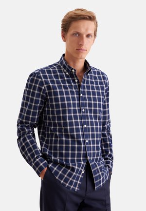 Homme portant une chemise à carreaux bleu marine et blanc à manches longues et un pantalon bleu marine, debout les mains dans les poches.