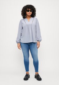 Gai pelēks garu piedurkņu blūze ar V-veida kakla izgriezumu, sacelto jostu un dekoratīvām šuvēm. Kombinēta ar ziliem šauriem džinsiem un masīvām melnām klumpēm.