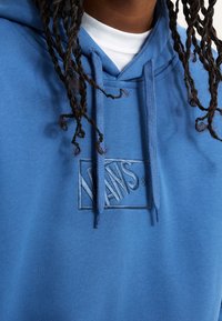 Personne aux cheveux tressés portant un sweat à capuche bleu Vans par-dessus un t-shirt blanc, mettant l'accent sur le logo du sweat et les cordons.