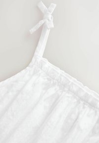 White cotton camisole featuring thin straps with knotted details and an elastic waist, showcasing a soft texture and light, airy design.