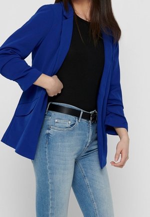 Kvinde iført en knaldblå blazer, sort skjorte, lyseblå jeans og sort bælte, med den ene hånd nær taljen og den anden afslappet.