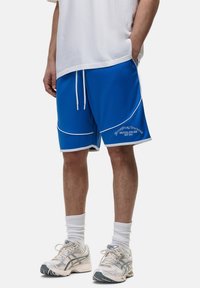 Pantaloni sportivi blu con piping bianco e un logo, realizzati in un tessuto leggero. Abbinati a scarpe da ginnastica grigie e calze bianche.