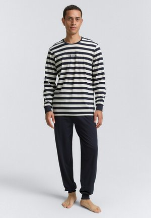 Ein Mann steht barfuß und trägt ein schwarz-weiß gestreiftes Langarmshirt sowie lockere schwarze Hosen vor einem schlichten Hintergrund.