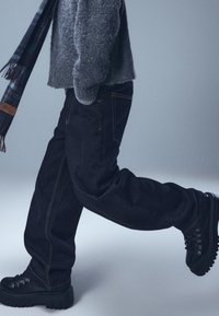 Crne traperice širokog kroja u paru s crnim čizmama s debelim potplatom. Oprema uključuje sivu teksturiranu majicu i kariranu šal.