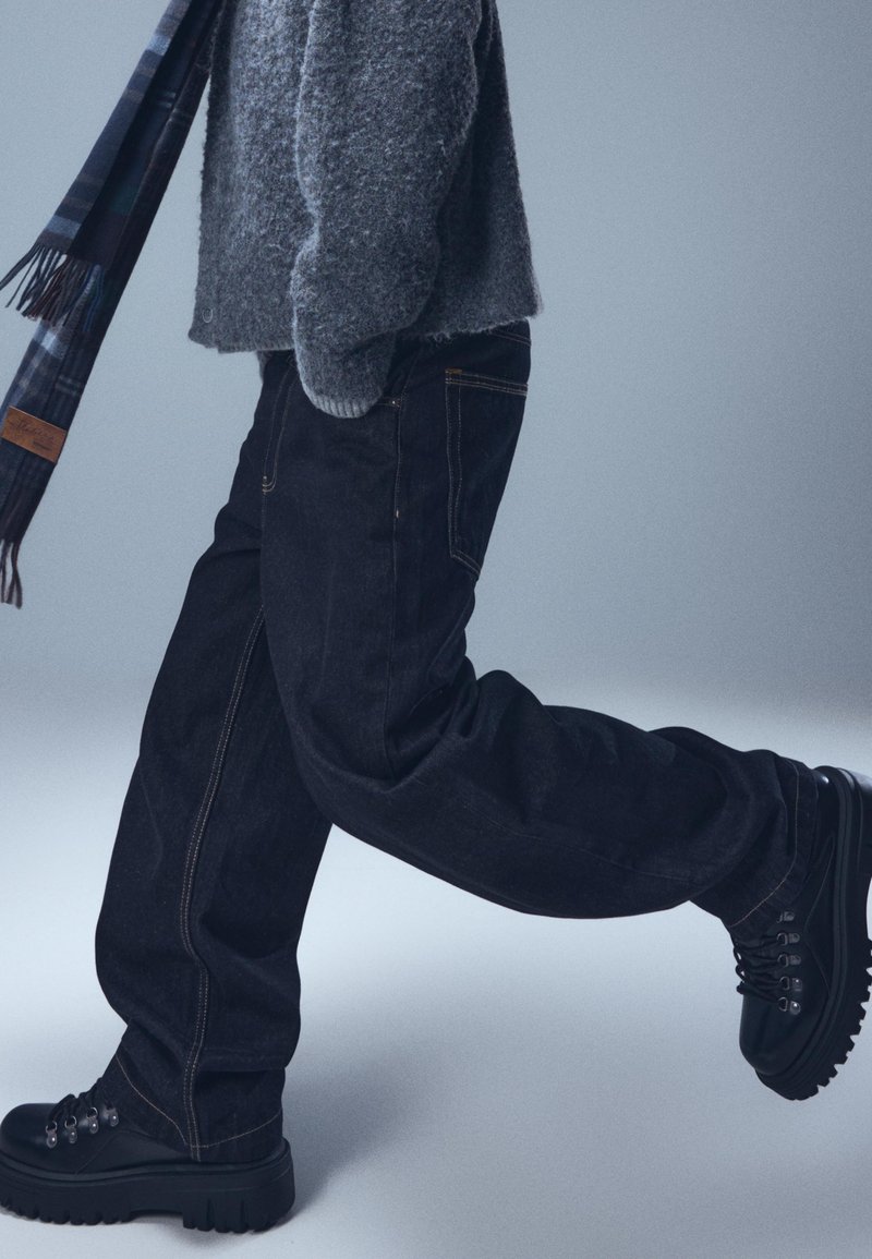 Crne traperice širokog kroja u paru s crnim čizmama s debelim potplatom. Oprema uključuje sivu teksturiranu majicu i kariranu šal.