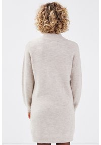 Femme aux cheveux blonds bouclés portant une robe-pull côtelée beige clair, debout de dos face au spectateur sur un fond uni.