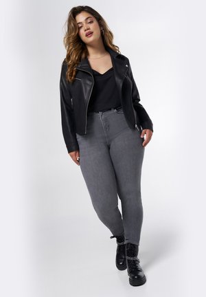 Zwarte leren cropped jack, zwarte top, hooggeknoopte grijze jeans en zwarte veterlaarzen met kettingdetails. Model poseert staand.