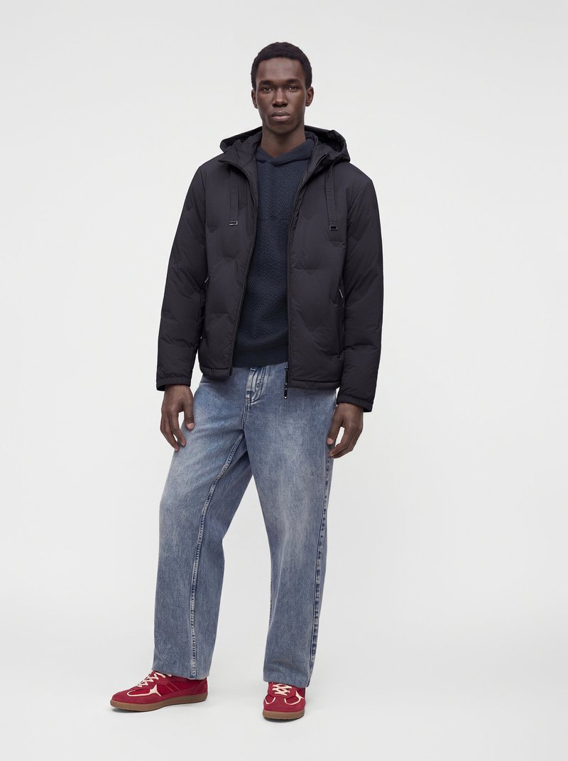 Jeune homme debout portant une veste noire à capuche, un pull foncé, un jean bleu clair ample et des baskets rouges sur un fond clair uni.