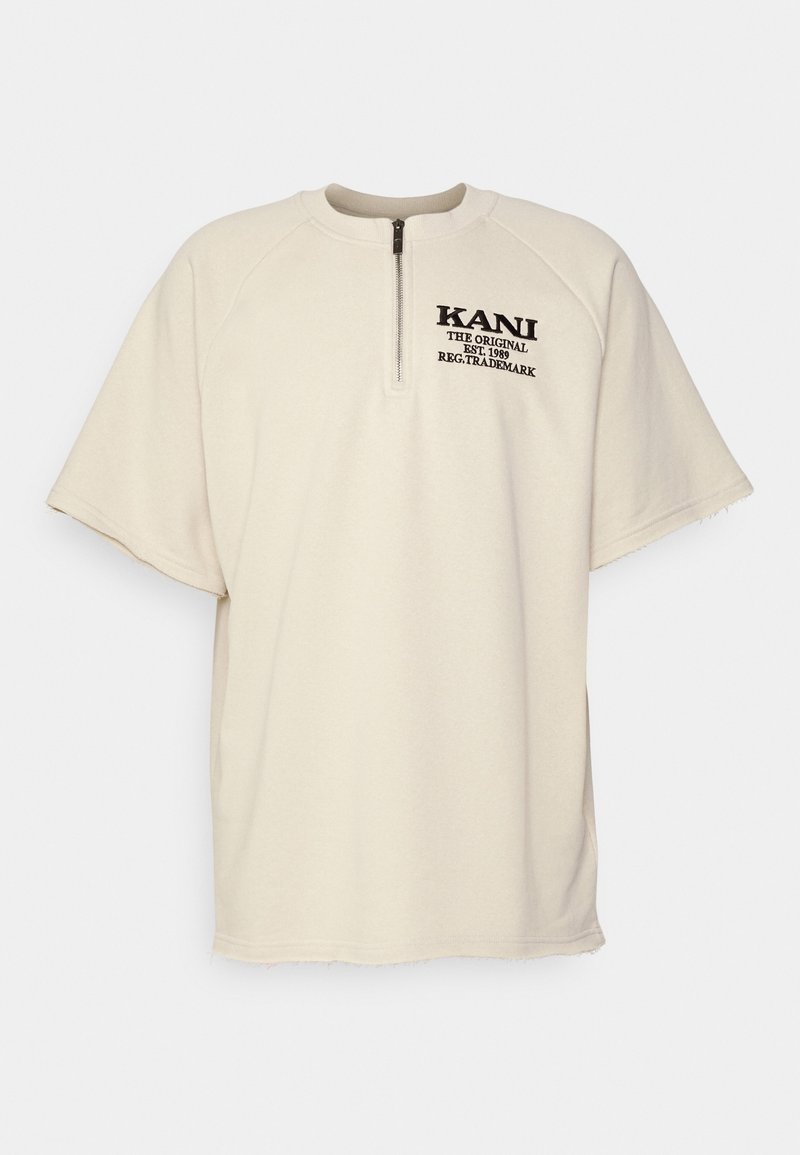 Pull à manches courtes beige avec fermeture éclair quart de longueur et texte noir « KANI The Original Est. 1989 Reg. Trademark » sur la poitrine gauche.