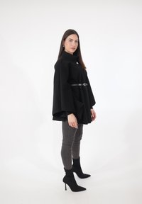 Sort uldponcho med høj krave, båret med bælte i taljen, over grå jeans og spidse sorte hæle. Glat tekstur, minimalistisk design.