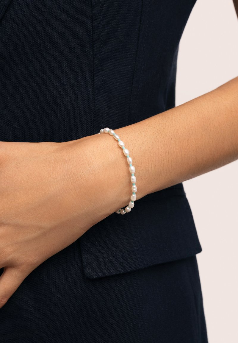 Bracciale di perle con piccole perle bianche rotonde, infilate insieme. Indossato su una mano con un tono di pelle chiaro, su uno sfondo di tessuto scuro.