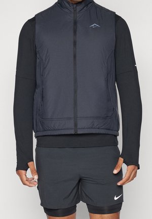 Mężczyzna ubrany w czarną kamizelkę bez rękawów zapinaną na zamek, na koszulce z długim rękawem z otworami na kciuki, czarne spodenki oraz czarne legginsy kompresyjne.