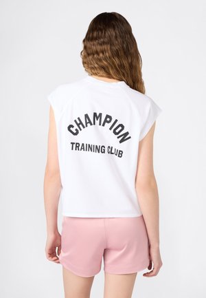 Mujer con cabello ondulado que lleva una camiseta blanca sin mangas con el texto "CHAMPION TRAINING CLUB" y pantalones cortos deportivos rosas, vista desde atrás.
