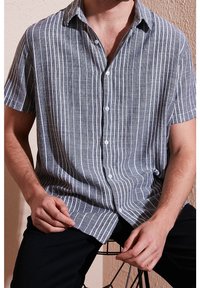Homme portant une chemise grise à manches courtes boutonnée avec des rayures blanches verticales, assis sur une chaise devant un fond uni.