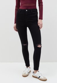 Jeans ajustados negros con detalles rasgados en las rodillas, cintura alta y textura de denim suave, combinados con zapatillas blancas y beiges.