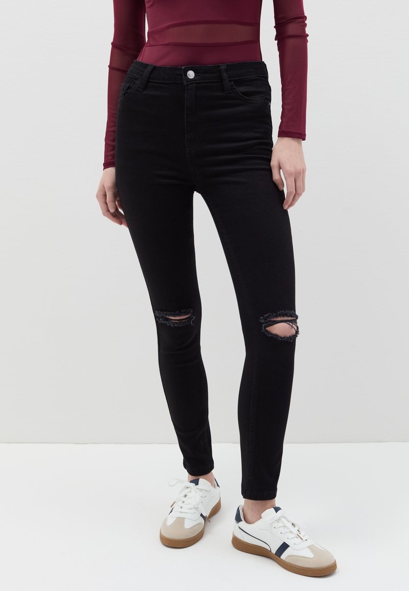 Jeans ajustados negros con detalles rasgados en las rodillas, cintura alta y textura de denim suave, combinados con zapatillas blancas y beiges.