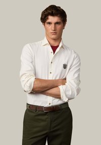 Camisa blanca de botón de manga larga con las mangas enrolladas, que presenta un parche de logo negro. Combinada con pantalones verde oscuro y un cinturón marrón.