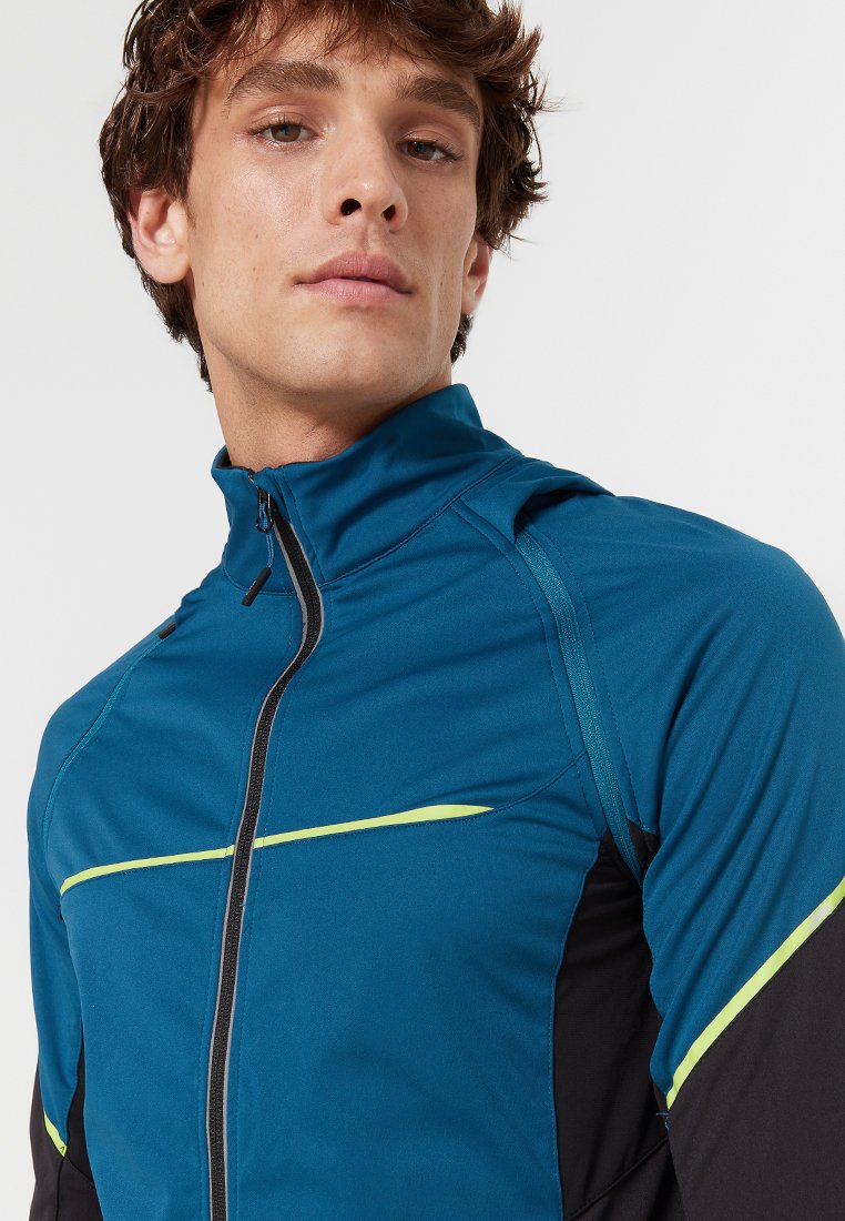Giacca sportiva in teal con accenti neri, dotata di un colletto alto, zip frontale e dettagli riflettenti gialli sul petto e sulle spalle.