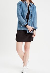 Veste en denim avec une teinte bleue, fermeture par boutons et deux poches avant ; portée sur une robe noire et associée à des baskets blanches.