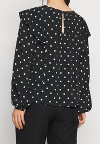 Blusa nera con pois bianchi, con maniche a volant e un dettaglio a occhiello sul retro. Tessuto morbido e vestibilità ampia.