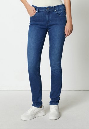 Jean slim fit en denim bleu porté avec des baskets blanches, modèle avec une main dans la poche, debout devant un fond clair uni.