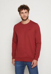 Roter Baumwoll-Sweatshirt mit langen Ärmeln und Rundhalsausschnitt. Verfügt über gerippte Bündchen und Saum, kleines Logo auf der Brust, kombiniert mit blauen Jeans.