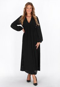 Rochie lungă neagră dintr-un material ușor, cu decolteu adânc în V, mâneci lungi bufante și talie strânsă. Asortată cu pantofi cu toc negri.