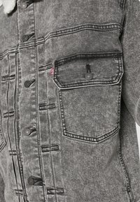 Grå denimjacka med en strukturerad yta, knäppning med knappar, dubbla bröstfickor och kontrasterande sömnadsdetaljer.