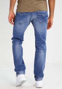 Jeans de denim azul para hombre con corte recto, detalles desgastados en los bolsillos y una textura suave. Llevados con zapatillas blancas.
