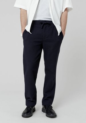 Pantalons en mélange de lin bleu marine avec ceinture élastique réglable, poches latérales et jambes fuselées, associés à un t-shirt gris clair et une chemise blanche.