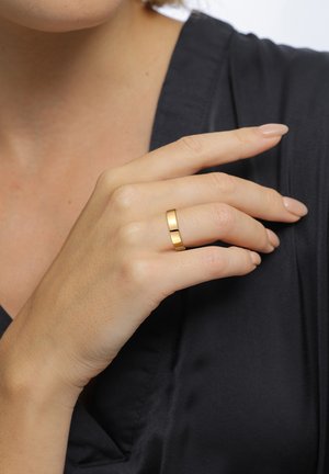 Gold metal ring with a flat surface and subtle cutout detail, worn on a hand with neutral nail polish, against a dark fabric background.