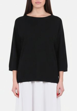 Donna che indossa un maglione nero a maniche lunghe e ampio e una gonna bianca, in piedi su uno sfondo bianco uniforme.